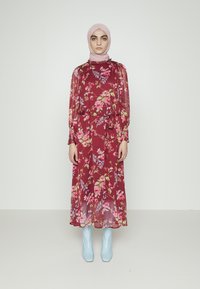Vestido floral vinho com sobreposição transparente, mangas longas e uma silhueta fluida. Combinado com botas ankle azuis claras. O hijab rosa completa o visual.