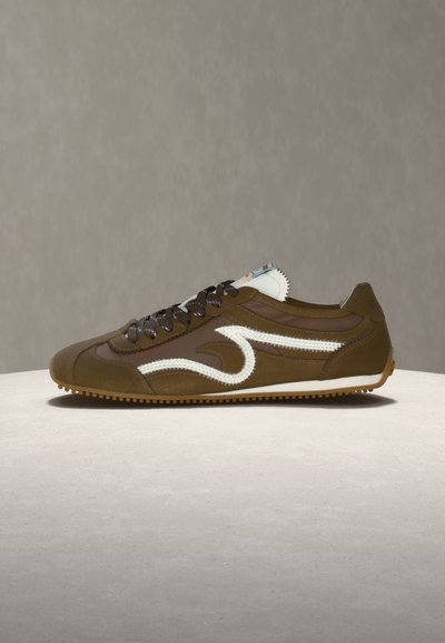 Καφέ αθλητικό παπούτσι χαμηλού ύψους με λευκό κυματιστό σχεδιασμό στο πλάι, μαύρα κορδόνια και ανοιχτή σόλα από καουτσούκ σε ουδέτερη επιφάνεια και φόντο.