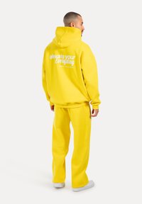 Hoodie gialla con testo sul retro e pantaloni joggers gialli abbinati. Entrambi gli articoli presentano un tessuto morbido e testurizzato e una vestibilità rilassata. Accenti bianchi visibili.