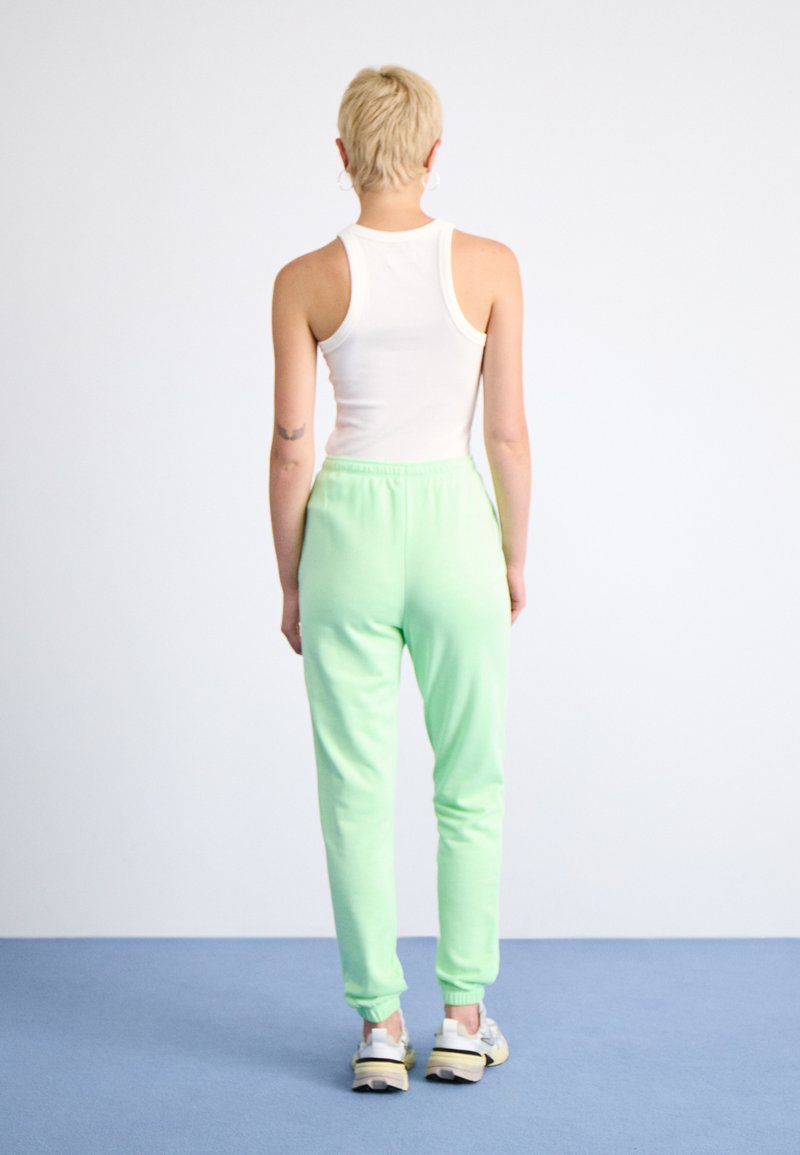 Hellgrüne Sweatpants mit elastischen Bündchen und Bund, kombiniert mit einem weißen ärmellosen Oberteil. Der Hintergrund ist schlicht.