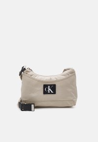 Bolso bandolera de nailon beige con forma curva, cremallera superior, correa ajustable y una etiqueta negra prominente con el logo "CK".