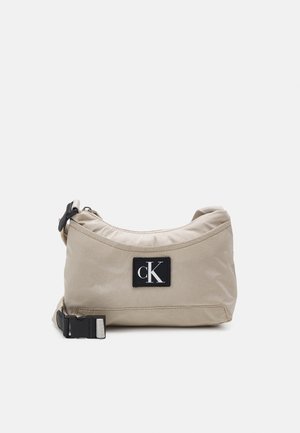 Beige nailonkassi, jossa on kaareva muoto, ylävetoketju, säädettävä hihna ja kuuluisa musta etiketti, jossa on "CK"-logo.