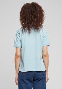 Lyseblå bomullspolo med korte ermer og rett nederkant, iført med blå denimbukser, viser bakvinkel og teksturert hår.