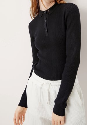Femme porte un polo noir côtelé ajusté à manches longues avec des boutons, associé à un pantalon blanc texturé avec cordon de serrage, sur un fond clair.