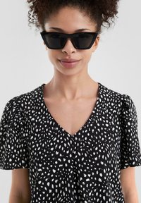 Schwarzes Kleid mit weißem Punktmuster, kurzen Puffärmeln und V-Ausschnitt. Modell trägt eckige schwarze Sonnenbrille vor einem neutralen Hintergrund.