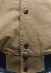 Nærbillede af beige jakke foran med fire sorte trykknapper og en ribbet mørkeblå manchet nederst.