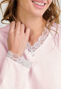 Mujer vestida con una blusa de color rosa claro con encaje blanco en el escote y los puños, sonriendo suavemente, con la mano tocando suavemente el cabello cerca de la cara.