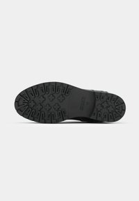 Semelle de chaussure noire avec des motifs de sculpture profonds comprenant des formes en croix et en crochet, présentée sur un fond uni gris clair.