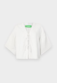 Hvit kort topp med V-hals, korte vide ermer, og snøring foran. Laget av pustende stoff. Grønn merkevarelabel synlig.