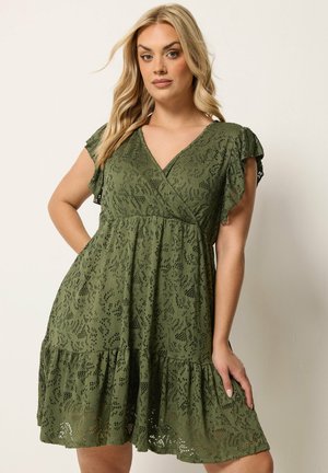 Femme blonde portant une robe en dentelle vert olive à manches courtes avec col en V et ourlet à volants superposés, debout devant un fond clair uni.