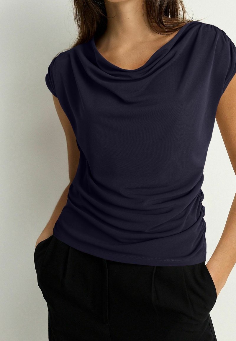 Top azul marino de manga corta con escote drapeado y fruncido lateral, hecho de un tejido suave y elástico. Combinado con pantalones negros.