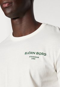 Tricou din bumbac de culoare albă murdară cu guler rotund. Prezintă text verde: "BJÖRN BORG STOCKHOLM 1956" în zona stângă a pieptului.