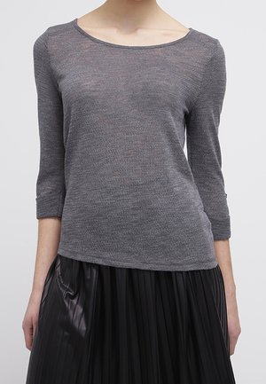 Personne portant un haut gris ajusté à manches longues avec un col dégagé et une jupe noire plissée sur un fond uni.