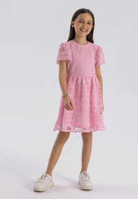 Rosa spetsklänning med korta puffärmar, prydd med intrikata blommönster och ett lager på lager- kjol. Bärs med vita sneakers.