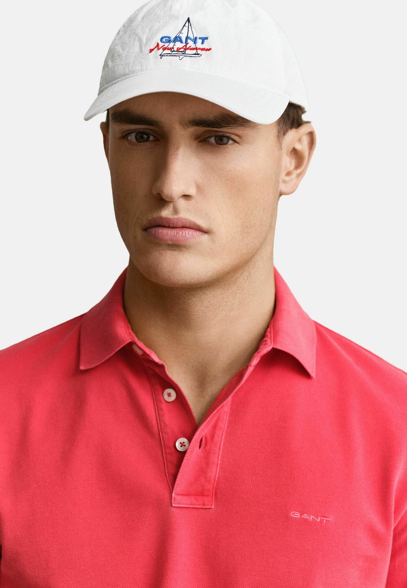 Mann mit weißer Kappe mit Segelboot-Logo und rotem Poloshirt mit GANT-Logo auf der Brust, neutraler Hintergrund.