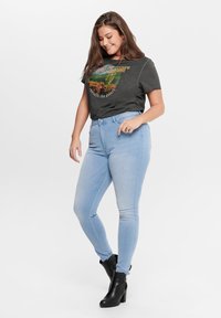 Γκρι γραφική μπλούζα με σχέδιο της ερήμου, συνδυασμένη με ανοιχτό μπλε στενά τζιν. Το μοντέλο φοράει μαύρες μπότες έως τον αστράγαλο και έχει έναν χαλαρό πόζα.