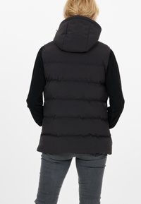 Sort puffer-vest med hætte, der har vandrette quiltede sektioner og en glat tekstur. Båret over en langærmet sort skjorte.