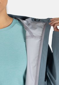Chaqueta resistente al agua en azul pizarra, con un forro interior en gris claro y cuello con cordón. Tejido texturizado con un acabado suave.