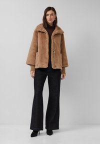Veste en fausse fourrure beige avec col, deux poches avant et fermeture à boutons. Associée à un pantalon large noir et un haut marron.
