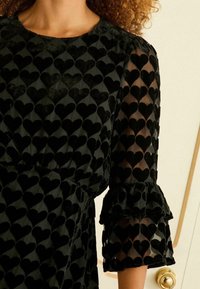 Čierne šaty s textúrovaným zamatovým korzetom so srdiečkovými vzormi a priesvitnými so špicatými rukávmi. Obohatené o volánik na manžetách.