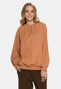 Przestronny sweter w kolorze rdzy z plisowanymi detalami przy dekolcie, długimi rękawami z elastycznymi mankietami oraz marszczonym dołem, wykonany z miękkiego materiału.