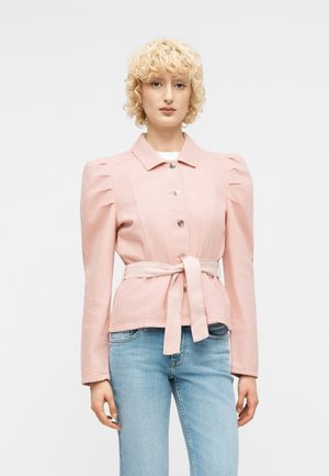 Kvinde iført en lyserød jakke med knapper, pufærmer og bånd i taljen, kombineret med lyseblå jeans, stående mod en hvid baggrund.