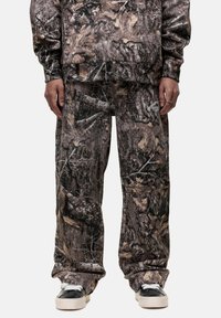 Camo-print sweatshirt and pants in brown, black, and earthy tones, featuring a relaxed fit and soft texture. Black footwear with white accents.