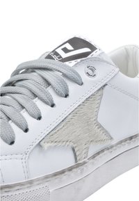 Białe skórzane sneakersy z teksturowanym szarym aplikacją w kształcie gwiazdy, szarymi sznurówkami i czarną metką z logo na języku. Gumowa podeszwa z niewielkimi śladami zużycia.