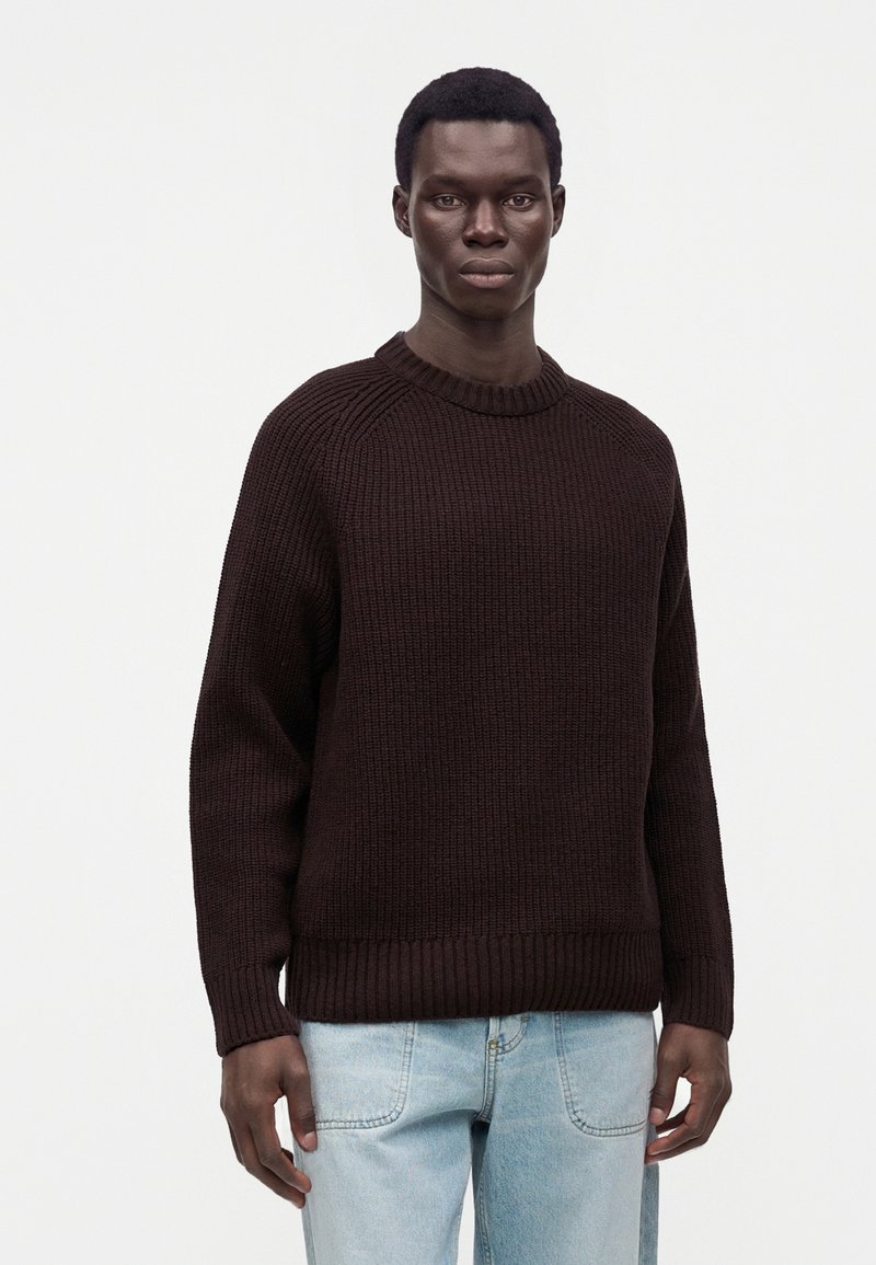 Homme portant un pull en maille côtelée marron foncé et un jean bleu clair, debout devant un fond clair uni.