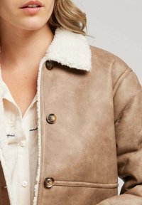 Veste en cuir marron avec un col en fausse fourrure blanche. Comprend des fermetures à boutons et une finition texturée. Pas de poches visibles.