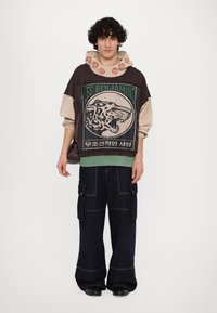 Sweatshirt surdimensionné en marron et beige avec un graphique de lion et des accents verts, associé à un pantalon cargo en denim foncé à jambe large.