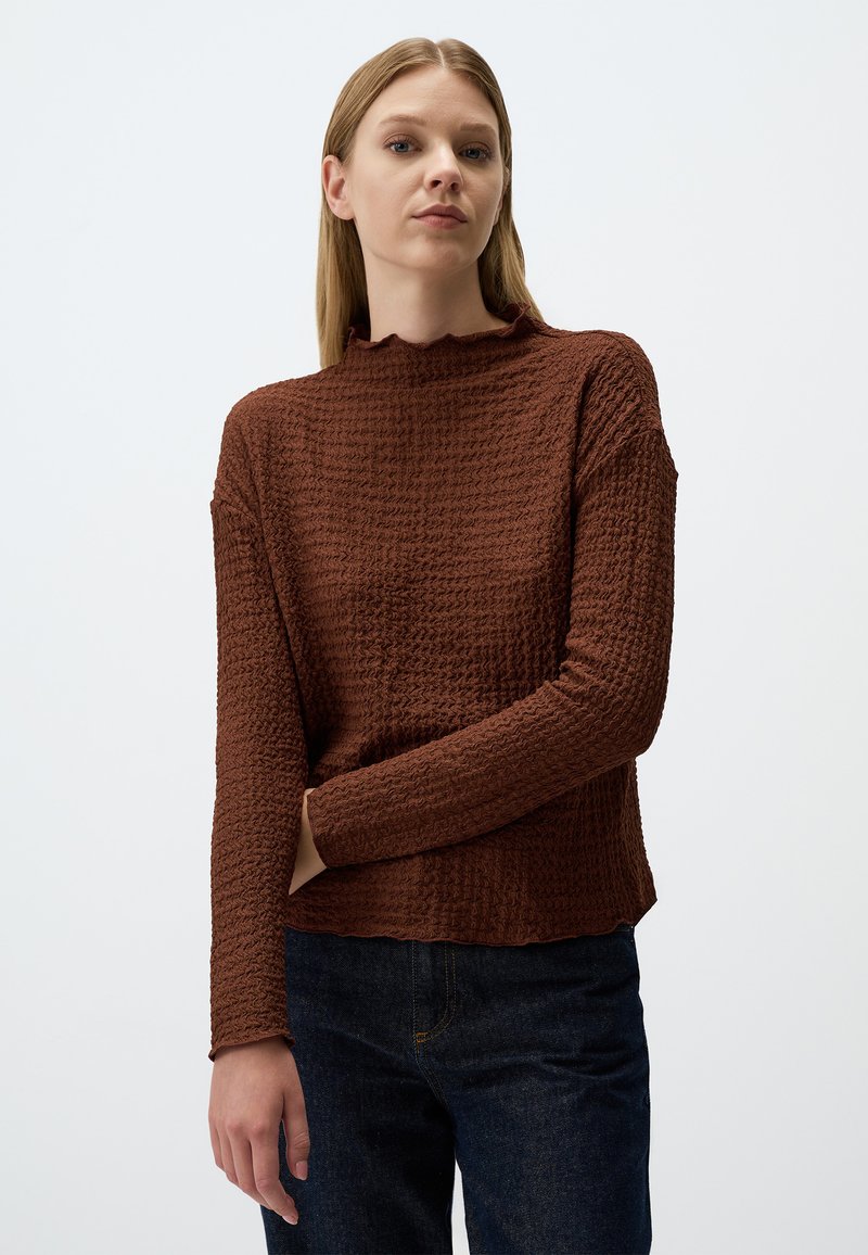 Un pull long à manches longues de couleur brune avec un col festonné, une coupe ample et des manches côtelées, associé à un jean en denim foncé.
