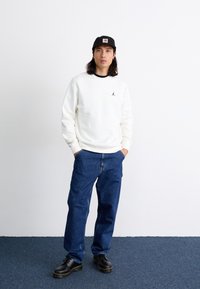 Hvid sweatshirts med sorte detaljer, prydet med et lille logo, parret med blå denim jeans og sorte snørebåndssko. Baggrunden er ensfarvet.