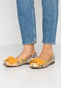 Baleriny z kolorową, drukowaną tkaniną na cholewce, z żółtą kokardką na czubku i elastyczną brązową podeszwą.