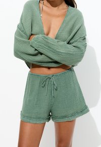 Femme portant un crop top vert à manches longues texturé et un short assorti à cordon, debout les bras croisés sur un fond blanc.