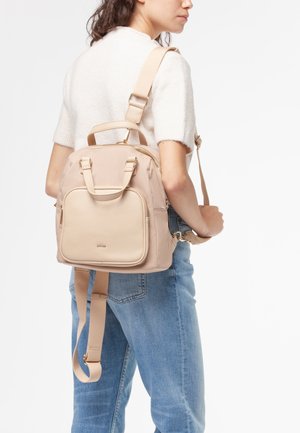 Donna che indossa uno zaino beige chiaro con tasca frontale e spallacci regolabili, abbinato a un maglione bianco a maniche corte e jeans blu.