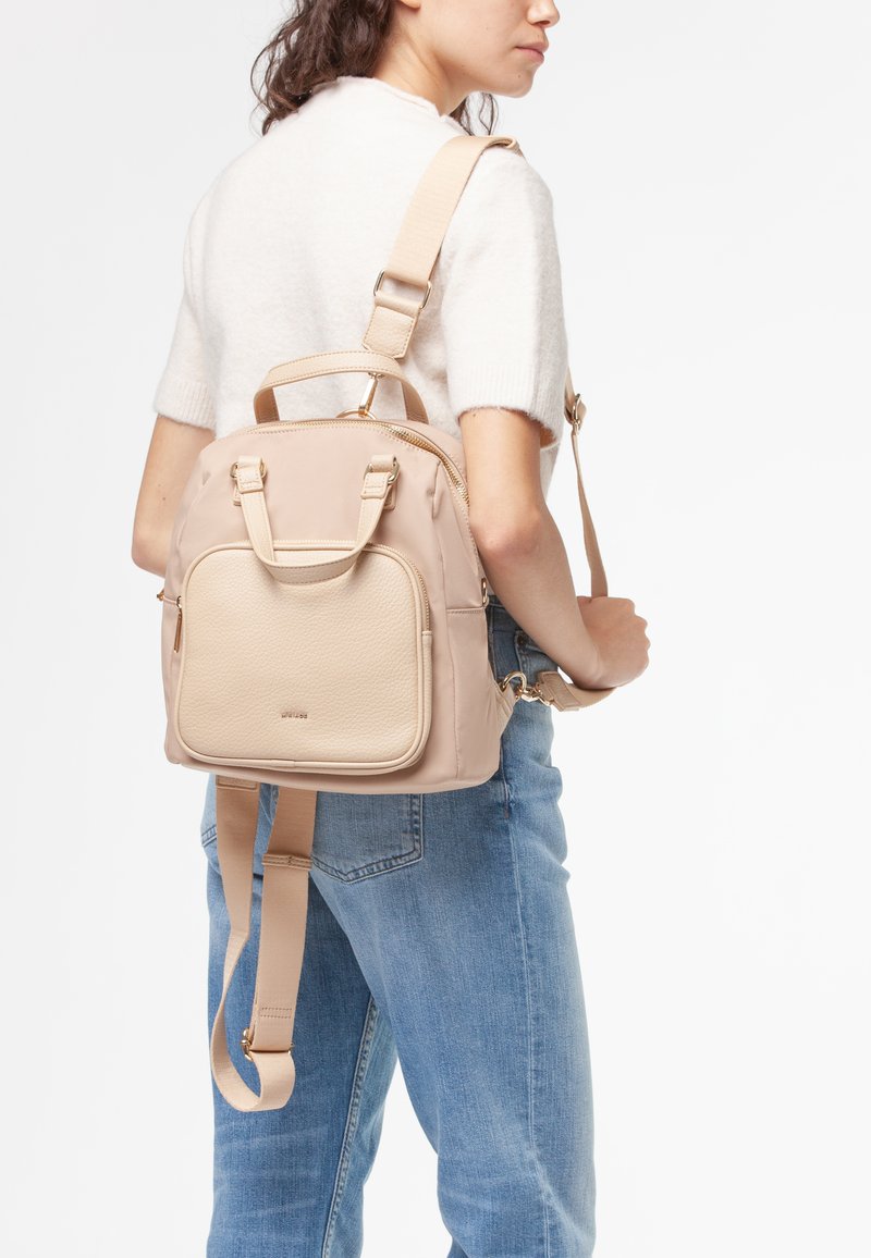 Mujer con una mochila beige claro que tiene un bolsillo frontal y tirantes ajustables, combinada con un suéter blanco de manga corta y jeans azules.