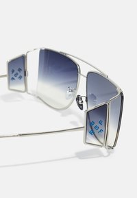 Occhiali da sole in metallo argento con montatura angolare, lenti grigie sfumate e branding blu sui pannelli laterali. Dettagli di design geometrico unici.