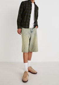 Grün-schwarz kariertes Hemd zum Knöpfen über einem weißen T-Shirt, kombiniert mit hellen Jeansshorts und beigen Sneakern mit orangefarbenen Akzenten.