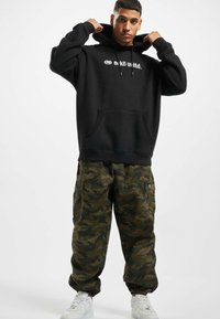 Svart hettegenser med hvitt logo på brystet, kengurulomme. Kamuflasje cargo-bukser med lommer og elastiske mansjetter. Hvite sneakers.