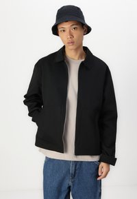 Veste zippée noire avec un col, deux poches à l'avant, portée sur une chemise gris clair. Arrière-plan blanc, avec un chapeau seau noir.