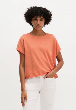 Woman with curly hair wearing a loose coral short-sleeve top and white pants, one hand in pocket, against plain white background.