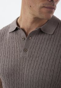 Lysebrun strikket polo-shirt med et tekstureret kabelmønster, der har en krave og to knapper ved halsen.