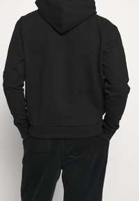 Felpa nera di cotone con cappuccio, orlo e polsini a costine. Indossata con pantaloni di velluto a coste scuri, che mostrano una finitura testurizzata sul tessuto.