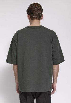 Uomo con capelli castani corti e ricci, che indossa una camicia verde scuro a maniche corte oversize e pantaloni neri, di spalle su uno sfondo neutro.