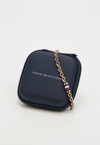 Tumed sinine ruudukujuline tõmblukuga kott, millel on "Tommy Hilfiger" logo ja kuldne kett, millel rippuv punane, valge ja sinine triibuline helmes.