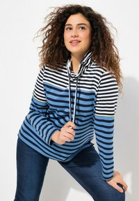 Blauer und weißer gestreifter Pullover mit einem Stehkragen, der eine Kordelzug und eine gelbe Akzentuierung am Ärmel hat. Getragen mit dunklen Jeans.