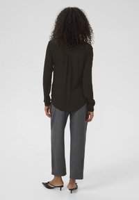 Dunkelgrünes Langarmshirt mit abgerundetem Saum; kombiniert mit grauen, weit geschnittenen Hosen und schwarzen Absatzmules. Glatte Stofftextur.