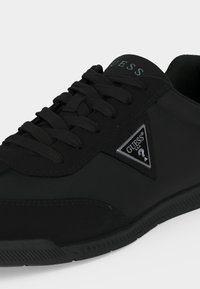 Baskets basses noires avec lacets noirs, semelle en caoutchouc et logo Guess triangulaire en métal sur le côté près des lacets.