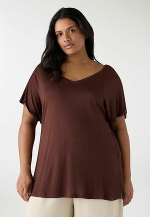 Donna con capelli scuri che indossa una maglietta marrone a scollo a V eampia e pantaloni beige chiaro, in piedi contro uno sfondo chiaro e semplice.